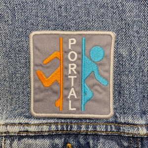 Può includere: Una toppa quadrata con la scritta "PORTAL" in lettere maiuscole bianche. La toppa presenta uno sfondo grigio con un bordo. Il design include figure arancioni e blu. La toppa è cucita su una giacca di jeans.