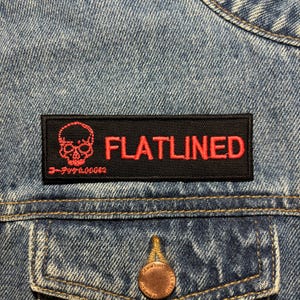 Może przedstawiać: Czarna łatka z czerwonym haftem "FLATLINED" i grafiką czaszki. Tekst "3-777-00003" znajduje się pod czaszką.