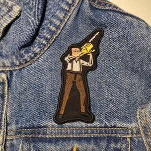 Puede incluir: Un parche bordado de una persona sosteniendo una motosierra amarilla. El parche es marrón y negro y está cosido en una chaqueta vaquera azul.