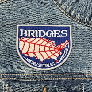 May include: A blue and white embroidered patch featuring the word "BRIDGES" above a red and white map of the United States. The patch is sewn onto a denim jacket and has the words "UNITED CITIES OF AMERICA" around the bottom edge.