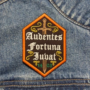 May include: A hexagonal patch with the Latin phrase "Audentes Fortuna Juvat" embroidered in white and orange on a black background. The patch features a sword and decorative flourishes, with the date "14 03" at the bottom. The patch is sewn onto a denim jacket.