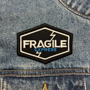 May include: An embroidered patch featuring the words "FRAGILE EXPRESS" in white and blue, with lightning bolt graphics. The patch is black with a white border, in a hexagon shape, and is sewn onto a blue denim jacket.