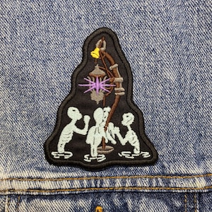 Op de afbeelding: Een geborduurde patch met drie skeletten in een donkere grot met een lantaarn die aan het plafond hangt. De lantaarn heeft een paars stervormig licht en een gele bel.