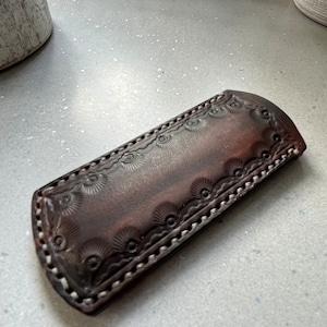 May include: A brown leather knife sheath with embossed floral patterns along the edges. The sheath is stitched with white thread and has a curved top. The leather has a distressed appearance.