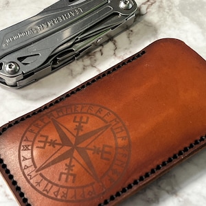 May include: A brown leather sheath featuring a circular compass design with runic symbols and black stitched edges, positioned next to a silver Leatherman Wingman multi-tool. The leather has a smooth finish and a deep tan colour with dark accents.