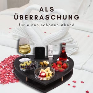 Könnte beinhalten: Ein schwarzes, herzförmiges Holz-Serviertablett mit Aussparungen für ein Weinglas, Smartphone, Fernbedienung und Flasche. Es enthält drei Glasschalen mit Weintrauben, Oliven, Käse und Erdbeeren. Der Text lautet: ALS ÜBERRASCHUNG für einen schönen Abend.