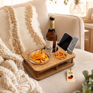 Könnte beinhalten: Ein braunes Couch-Tablett aus Holz mit einer runden Aussparung für eine Flasche, zwei Schlitzen für Smartphone und Fernbedienung sowie zwei Glasschalen mit Snacks. Ein kleines rechteckiges Etikett mit Herz und deutscher Flagge ist befestigt.