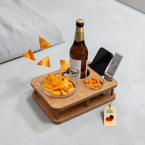 Puede incluir: Una bandeja de baño de madera con una botella de cerveza, aperitivos, un teléfono y un mando a distancia. La bandeja tiene un cuenco de vidrio transparente con totopos y un cuenco pequeño con frutos secos. Una etiqueta con un corazón y la bandera alemana cuelga de la bandeja.