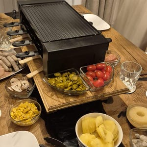 Könnte beinhalten: Ein schwarzer Raclette-Grill mit geriffelter Kochfläche steht auf einem Holztisch, umgeben von Schalen mit Zutaten wie Tomaten, Mais und Kartoffeln. Eine gemütliche Atmosphäre für ein Essen.