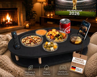 May include: A black oval couch bar with light wood legs, featuring cutouts for a remote control, a soda can, and a smartphone. It holds three glass bowls containing mixed nuts, chips, and olives with cheese, plus a glass with a lemon slice.