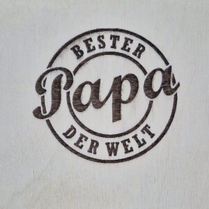 May include: A light-colored wooden surface with a dark brown circular design. The design features the words "BESTER Papa DER WELT" in a stylized font, with "Papa" being the most prominent word.
