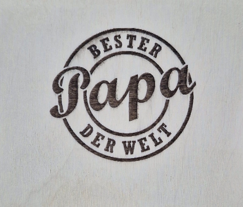 May include: A light-colored wooden surface with a dark brown circular design. The design features the words "BESTER Papa DER WELT" in a stylized font, with "Papa" being the most prominent word.