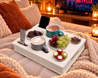 May include: A light-colored wooden serving tray with dedicated slots for a smartphone and remote control. The tray holds two mugs with coffee, a bowl of chocolate cookies, a bowl of green and red grapes, a small bowl of chocolate, and stuffed peppers.