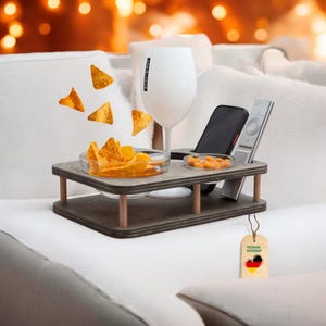 Könnte beinhalten: Ein Couchtablett aus Holz mit zwei Ebenen, mit einem Glas für Getränke, einer Schüssel für Snacks, einem Telefonhalter und einem Fernbedienungshalter. Das Tablett befindet sich auf einem weißen Sofa mit einem Etikett, auf dem "Premium Couchbar" steht.