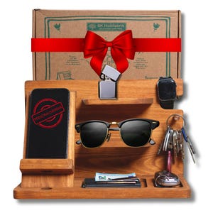 Könnte beinhalten: Ein Holz-Schreibtisch-Organizer mit Telefon, Sonnenbrille, Uhr, Schlüsseln, Geldbörse und Feuerzeug. Auf dem Telefon steht das Wort "PERSONALISIERBAR" in einem roten Stempel. Im Hintergrund eine Geschenkbox mit roter Schleife.