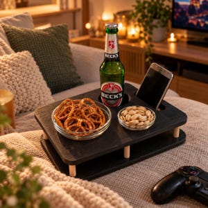 Könnte beinhalten: Ein schwarzes Snack-Tablett aus Holz mit vier hellen, zylindrischen Beinen, ausgestattet mit einer runden Aussparung für eine grüne Bierflasche, einem rechteckigen Schlitz für ein Smartphone sowie zwei Glasschalen mit Brezeln und Pistazien.