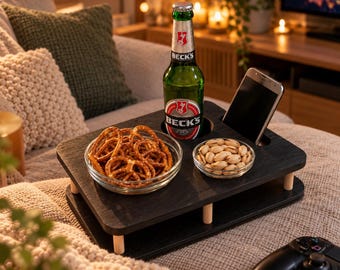 May include: A black wooden snack tray with four light-colored cylindrical legs, featuring a circular cutout for a green glass beer bottle, a rectangular slot for a smartphone, and two clear glass bowls containing pretzels and shelled pistachios.