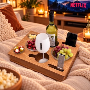 Könnte beinhalten: Ein rechteckiges Snack-Tablett aus Holz mit kreisförmigen Aussparungen für ein Weißweinglas und eine Weinflasche sowie Schlitzen für ein Smartphone und eine Fernbedienung. Drei Glasschalen enthalten Kirschen, Weintrauben, Käse und gefüllte Paprika.