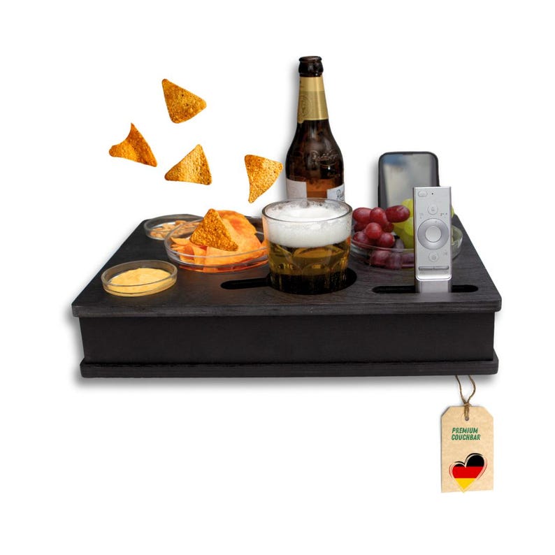 May include: A black wooden couch arm tray with cutouts for a beer glass, a bottle, and a remote control. The tray holds snacks like chips and dips, a beer, a phone, and grapes. A tag with a heart and the German flag hangs from the tray.