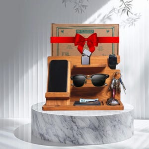 Könnte beinhalten: Ein hölzerner Schreibtisch-Organizer mit Telefonhalter, Sonnenbrille, Uhr, Schlüsseln und Feuerzeug. Eine Geschenkbox mit roter Schleife liegt obenauf. Der Organizer steht auf einem Marmorsockel.