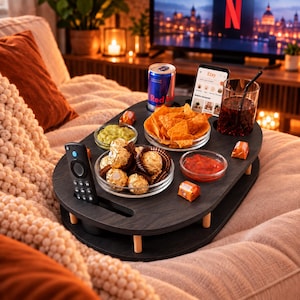 Könnte beinhalten: Ein ovales Couch-Tablett aus dunklem Holz mit kurzen Beinen und integrierten Schlitzen für eine Fernbedienung, eine Getränkedose und ein Smartphone. Das Tablett enthält eine Schale mit Tortilla-Chips, Guacamole, Salsa und goldverpackten Pralinen.