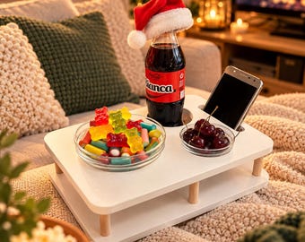 May include: A white wooden snack tray with rounded corners and four short legs. The tray holds a glass bottle of soda wearing a small Santa hat, a glass bowl filled with colorful gummy candies, a small bowl of dark cherries, and a smartphone.
