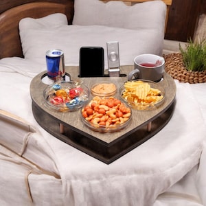 Puede incluir: Una bandeja de cama de madera en forma de corazón, gris, con compartimentos para aperitivos, una bebida, un teléfono y un mando a distancia. La bandeja contiene una lata de Red Bull, una taza y varios cuencos de cristal pequeños llenos de aperitivos.