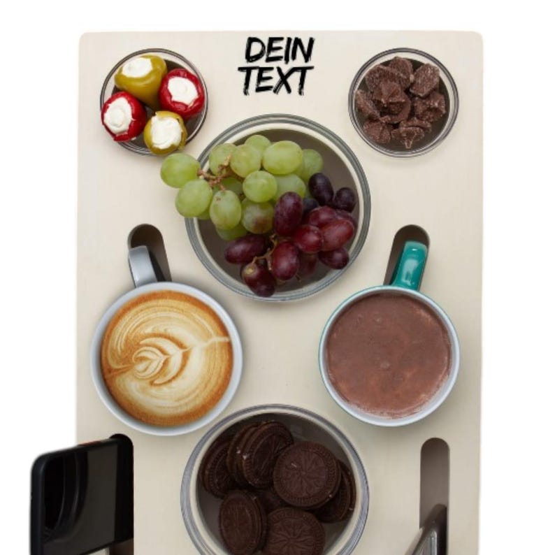 May include: A wooden serving board with various food items arranged in small bowls. Includes grapes, cookies, coffee, and stuffed peppers. The text "DEIN TEXT" is printed at the top. The board has a modern design.