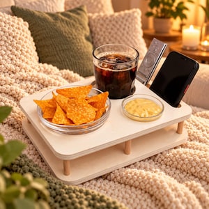 Puede incluir: Una bandeja de madera clara para aperitivos con un hueco circular para un vaso de bebida fría oscura, una ranura rectangular para un smartphone, una ranura para un mando a distancia y dos huecos para un cuenco de chips de tortilla y una salsa amarilla.