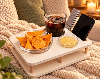 May include: A light-colored wooden snack tray featuring a circular cutout for a glass of dark iced beverage, a rectangular slot for a smartphone, a vertical slot for a remote control, and two recessed areas holding a glass bowl of tortilla chips and a small dish of yellow dip.
