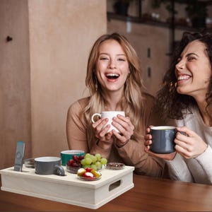 May include: A light-colored wooden serving tray with a handle, holding a gray mug, a teal mug, and small dishes of fruit and snacks. Two women are smiling and holding mugs. A remote control is also on the tray.