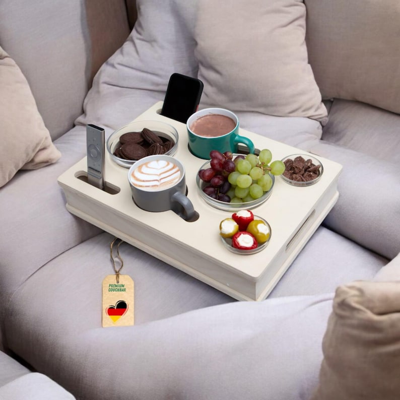 May include: A cream-colored wooden tray with a variety of snacks and drinks. The tray holds two mugs, a bowl of cookies, a bowl of chocolate, a bowl of grapes, and a bowl of stuffed peppers. A remote control and a smartphone are also on the tray.