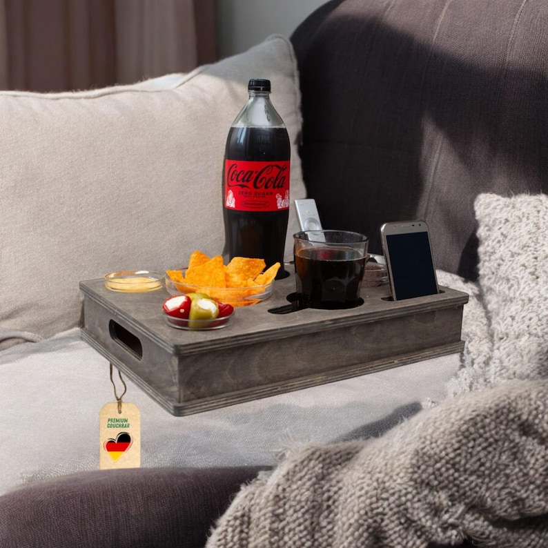 May include: A gray wooden couch tray with a bottle of Coca-Cola, a glass of soda, snacks, and a smartphone. The tray has a built-in phone holder and cup holder. A tag with the German flag hangs from the tray.