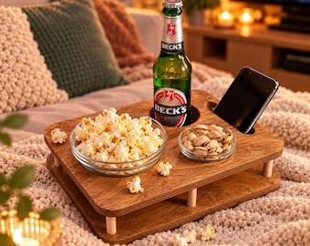 May include: A rectangular wooden snack tray with rounded corners and light-colored cylindrical legs. The tray features circular cutouts holding a green glass bottle of Beck's beer and a black smartphone, alongside two glass bowls containing popcorn and pistachios.