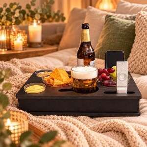 Könnte beinhalten: Ein schwarzes Holztablett mit integrierten runden Aussparungen für eine Bierflasche und ein Bierglas sowie rechteckigen Schlitzen für ein Smartphone und eine Fernbedienung, ergänzt durch kleine Glasschalen mit Tortilla-Chips, Dip und Weintrauben.