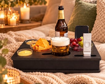 May include: A black wooden snack tray featuring integrated circular cutouts for a beer bottle and a glass of beer, along with rectangular slots for a smartphone and a remote control, plus small glass dishes containing tortilla chips, dip, and grapes.
