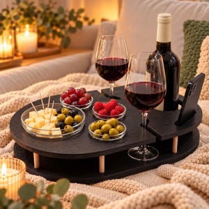 Puede incluir: Una bandeja de servicio de madera negra con patas cortas, que cuenta con ranuras integradas para dos copas de vino, una botella de vino tinto y un teléfono inteligente. La bandeja sostiene cuatro cuencos de cristal con dados de queso, aceitunas verdes, uvas rojas y frambuesas frescas.