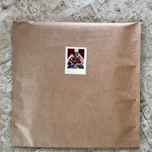 May include: A large, rectangular package wrapped in brown paper. A small sticker with an illustration of a person and flowers is affixed to the front. The package rests on a patterned, neutral-toned rug.