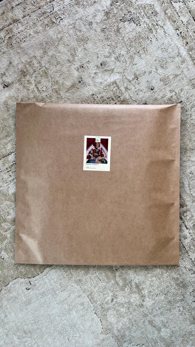 May include: A large, rectangular package wrapped in brown paper. A small sticker with an illustration of a person and flowers is affixed to the front. The package rests on a patterned, neutral-toned rug.