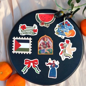 May include: A selection of stickers featuring Palestinian motifs, such as a watermelon, a flag-themed stamp, and floral illustrations. The stickers are displayed on a dark blue circular surface.