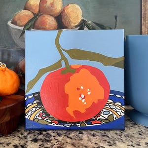 May include: A square canvas painting of a red and orange fruit with green leaves against a light blue background. The fruit sits on a blue plate with a decorative pattern. A painting of oranges is in the background.