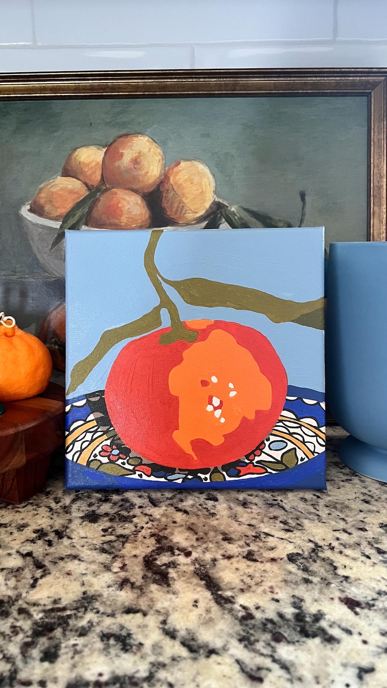 May include: A square canvas painting of a red and orange fruit with green leaves against a light blue background. The fruit sits on a blue plate with a decorative pattern. A painting of oranges is in the background.