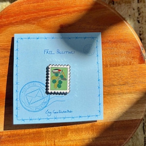 May include: A light blue card with the text "Free Palestine!" and a stamp featuring olive branches and the Palestinian flag. The card also has a post office stamp and the Etsy shop name "Etsy Gaza Palestine Studio."