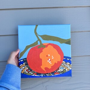May include: An acrylic painting of a red tomato with an orange section, set against a light blue background. The tomato rests on a patterned plate with a blue border. Green leaves and a stem extend from the tomato.