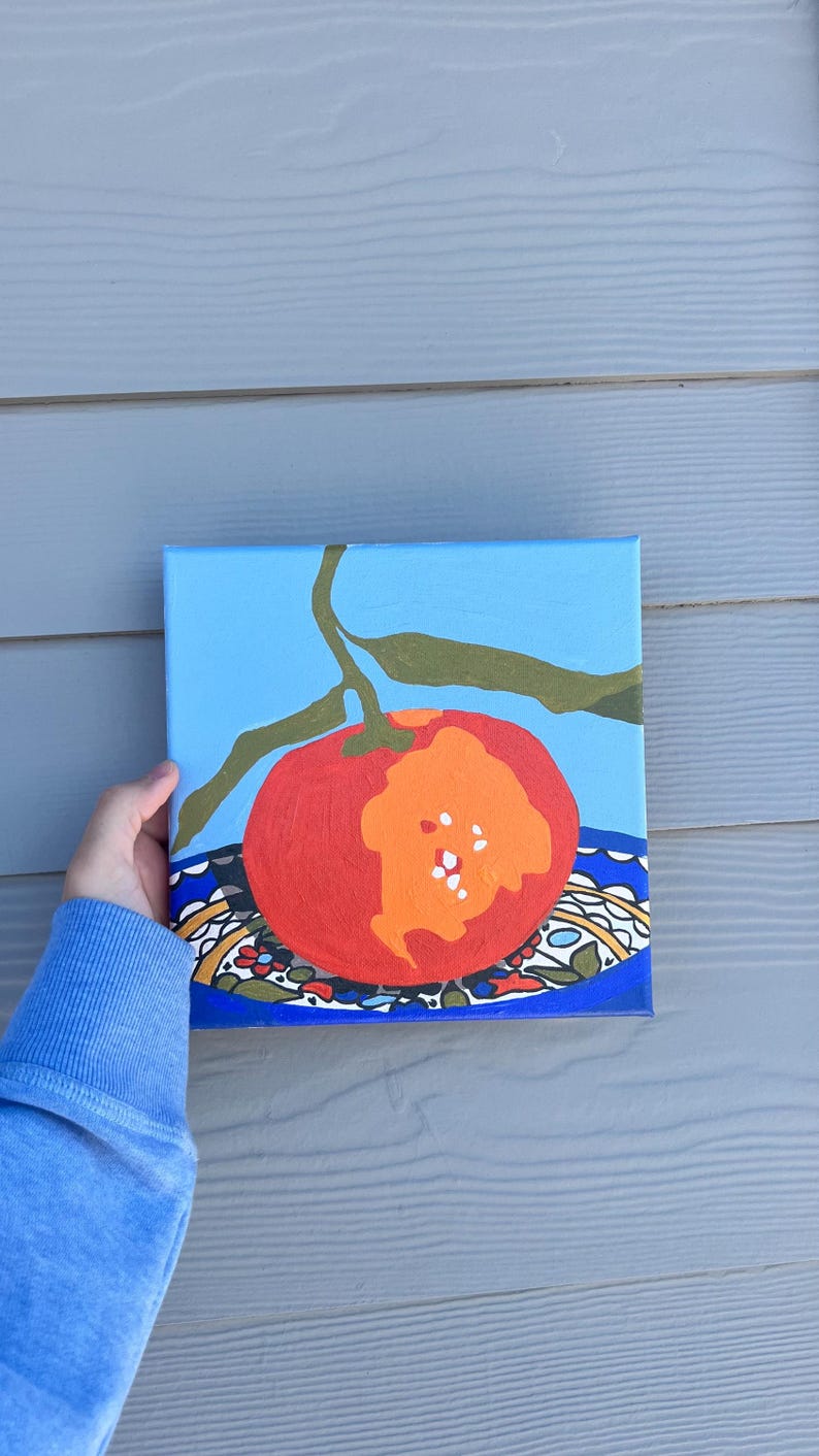 May include: An acrylic painting of a red tomato with an orange section, set against a light blue background. The tomato rests on a patterned plate with a blue border. Green leaves and a stem extend from the tomato.