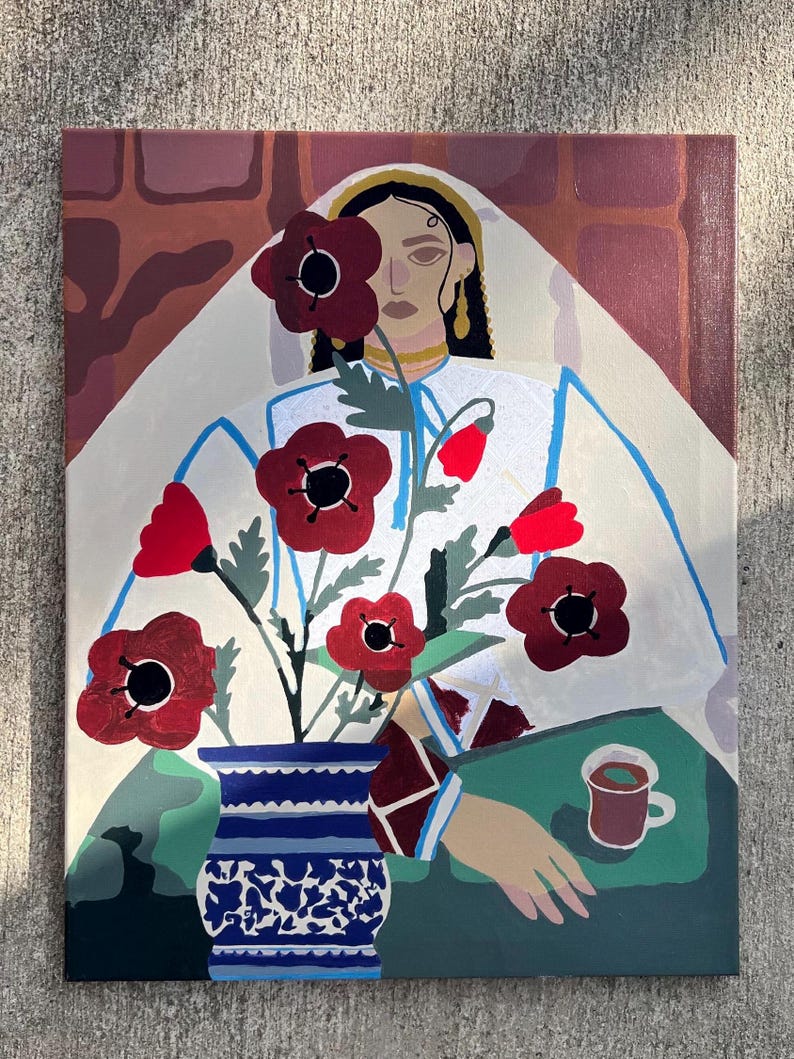 May include: An original painting featuring a woman in white attire with red poppies in a blue and white vase. A cup of coffee sits on a green surface. The background includes a brick-like pattern.
