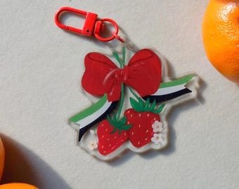 Könnte beinhalten: Ein klarer Acryl-Schlüsselanhänger mit einer roten Schleife, zwei Erdbeeren und einem grün-schwarz-weißen Band. Der Schlüsselanhänger hat einen roten Verschluss. Das Design befindet sich auf einem hellen Hintergrund mit Orangen.