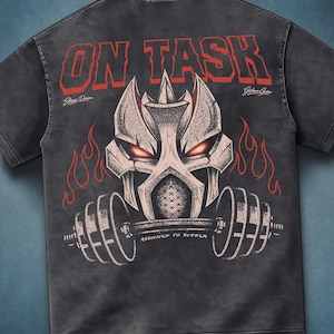 May include: Dark gray t-shirt with a distressed graphic. The graphic features a stylized helmet with glowing red eyes, a barbell, and flames. The text "ON TASK" is above the helmet, and "ASSIGNED TO SUFFER" is below.