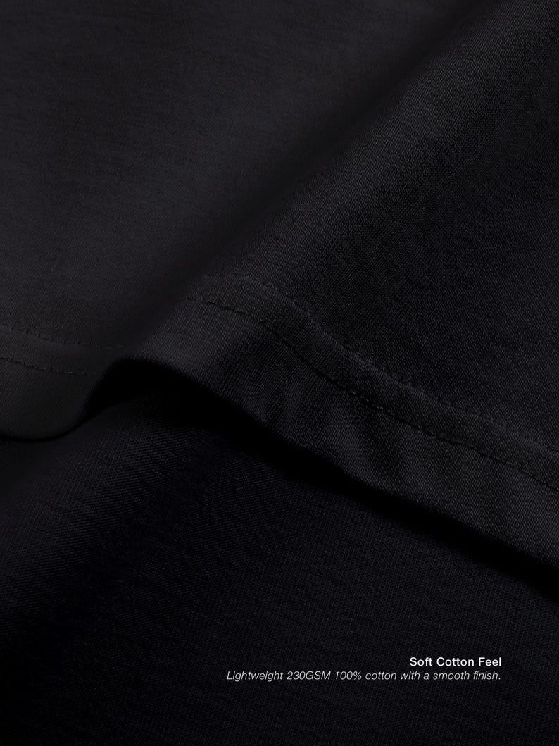 Op de afbeelding: Close-up van zwarte t-shirtstof, met de textuur en stiksels. De tekst onderaan luidt: "Soft Cotton Feel Lightweight 230GSM 100% cotton with a smooth finish."