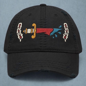 Puede incluir: Gorra de béisbol negra con visera desgastada. La gorra presenta un diseño bordado de una daga con detalles en rojo, azul y dorado. El diseño incluye elementos estilizados a ambos lados de la daga.